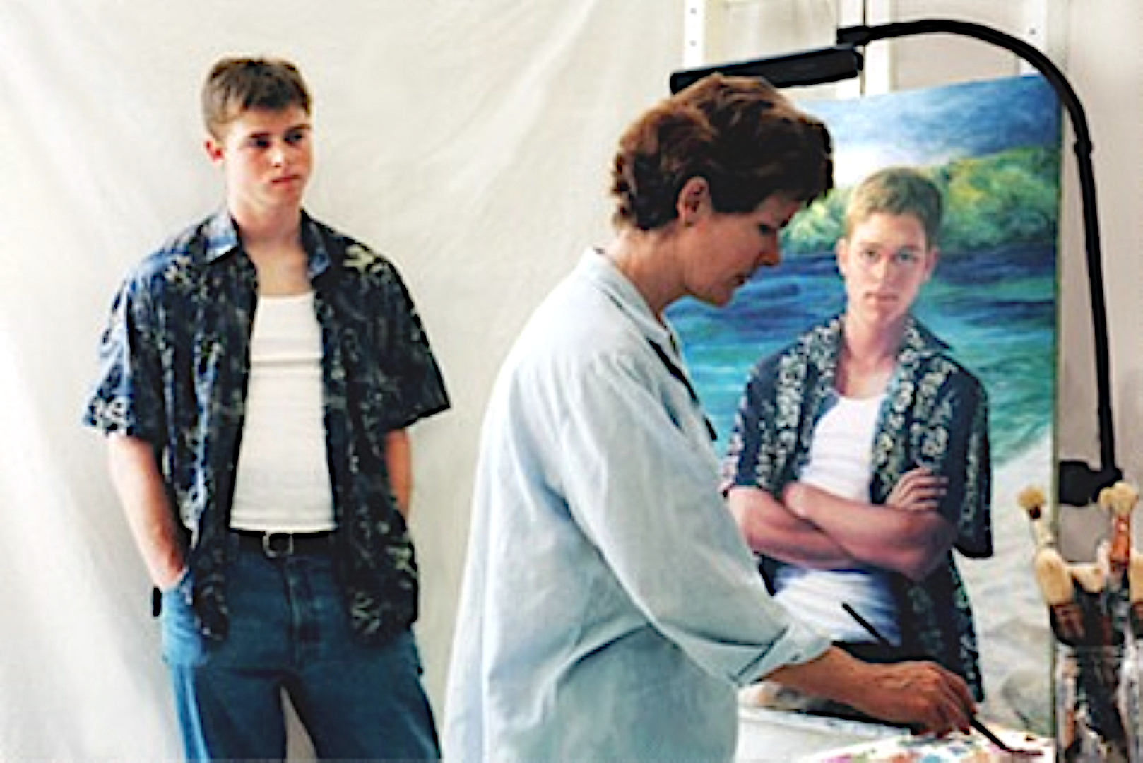 Photo of woman standing at easel painting portrait of teenage boy posing in hawaiian shirt.