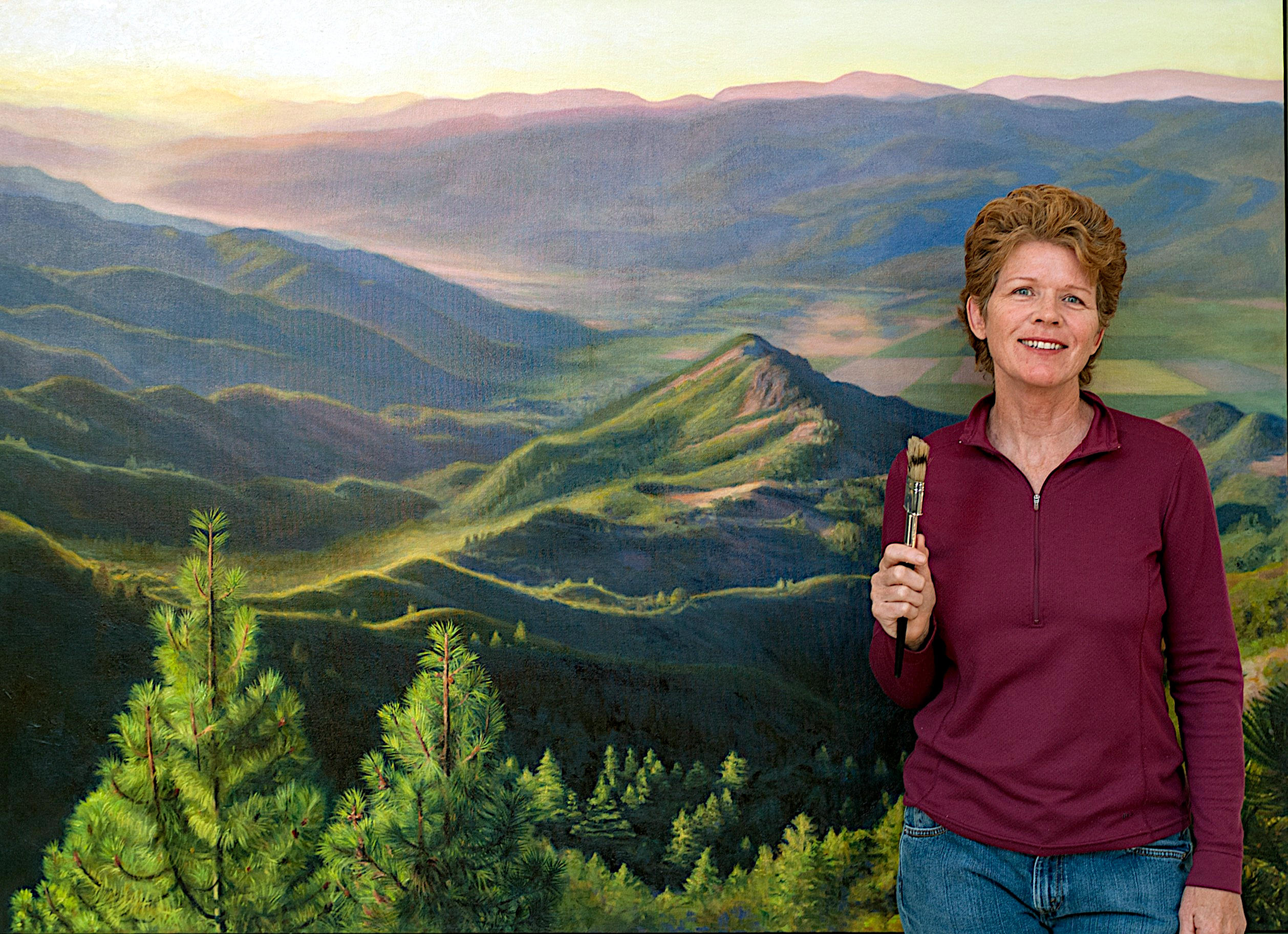 Photo of smiling woman holding paint brush and standing in front of painting.
