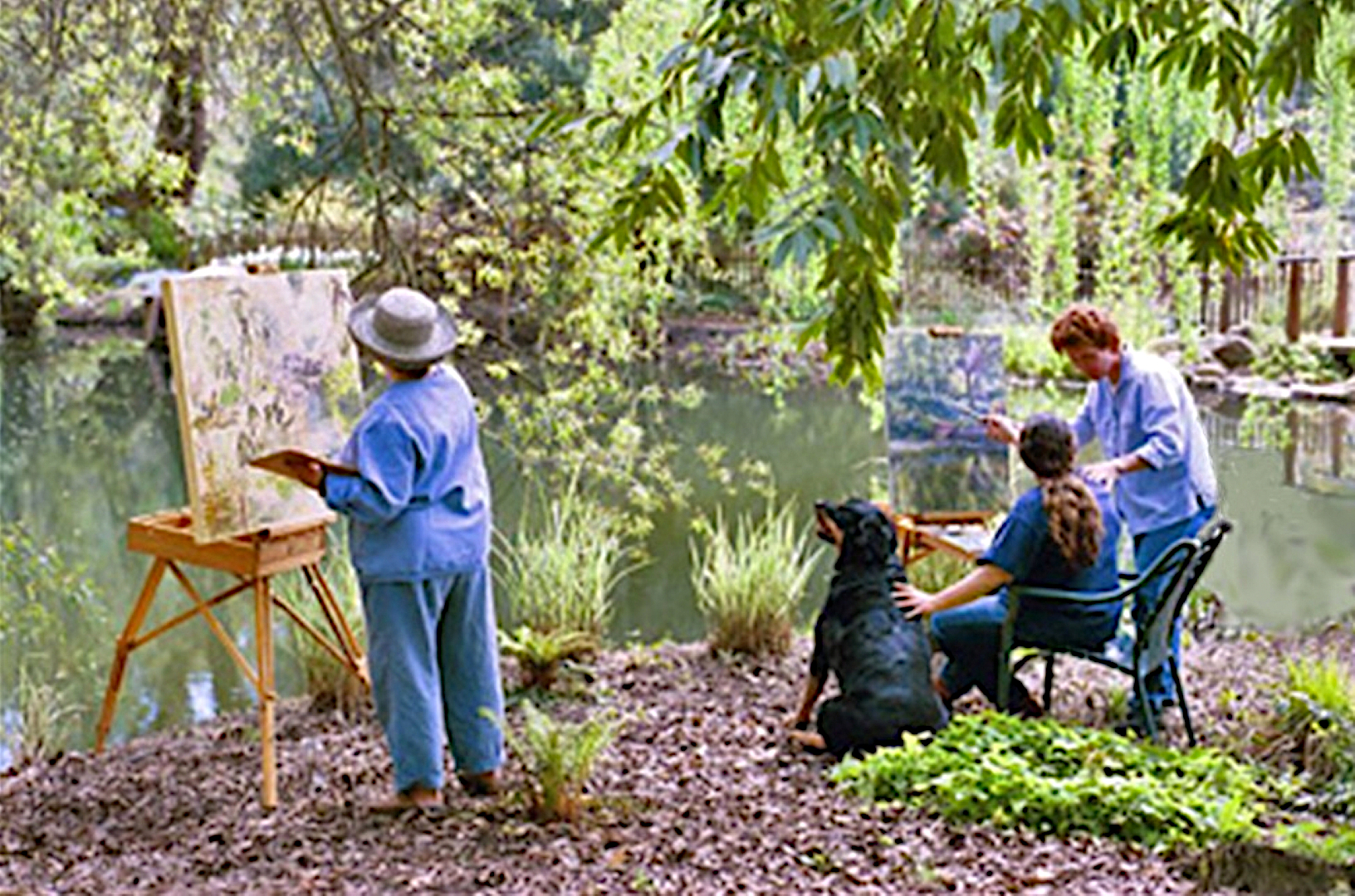 Photo of two woman sitting at edge of garden pond painting with easels, and third woman giving instructions.
